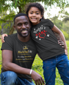 Man and child in matching 'Daddy's Girl' shirts posing outdoors on grass.