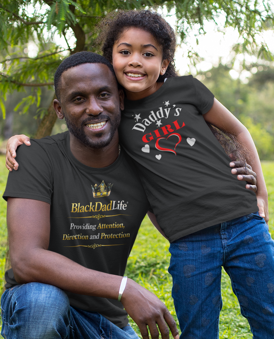 Man and child in matching 'Daddy's Girl' shirts posing outdoors on grass.
