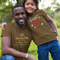 Man and child wearing matching brown t-shirts in a grassy outdoor setting