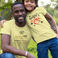 Man and young girl wearing matching yellow t-shirts in a grassy outdoor setting