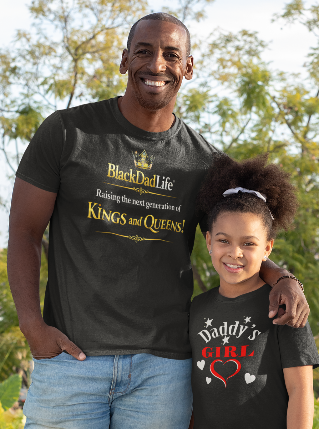 Man and young girl standing outdoors wearing matching black t-shirts with text.