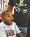 Child in a toy store with a surprised expression, wearing a 'Black Dad Life' t-shirt.