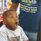 Child in a toy store with a surprised expression, wearing a 'Black Dad Life' t-shirt.