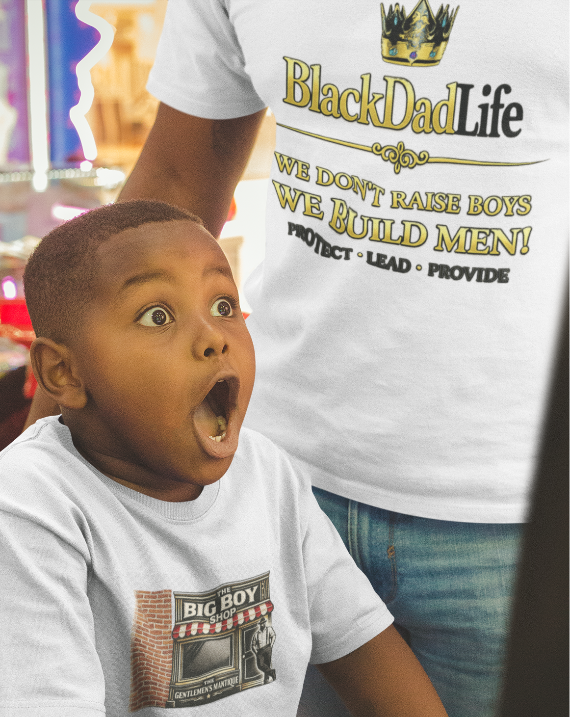 Child in a toy store wearing a 'Big Boy' t-shirt with an adult in a 'Black Dad Life' t-shirt in the background.