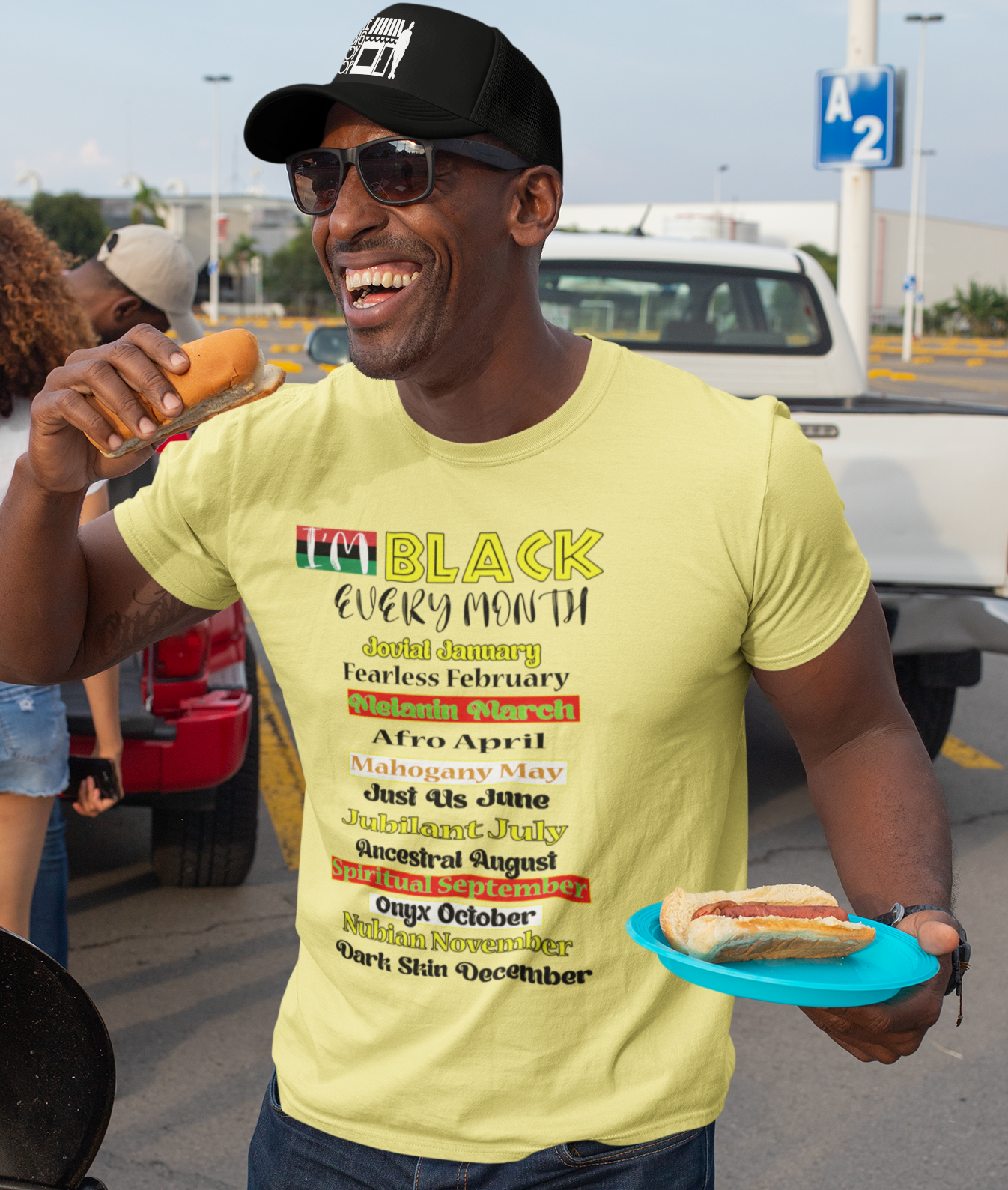 Man wearing a yellow t-shirt with 'Black History Month' text, holding a hot dog, standing outdoors.