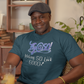 Man wearing a blue t-shirt with text, holding a glass of orange juice in a room with shelves and plants.