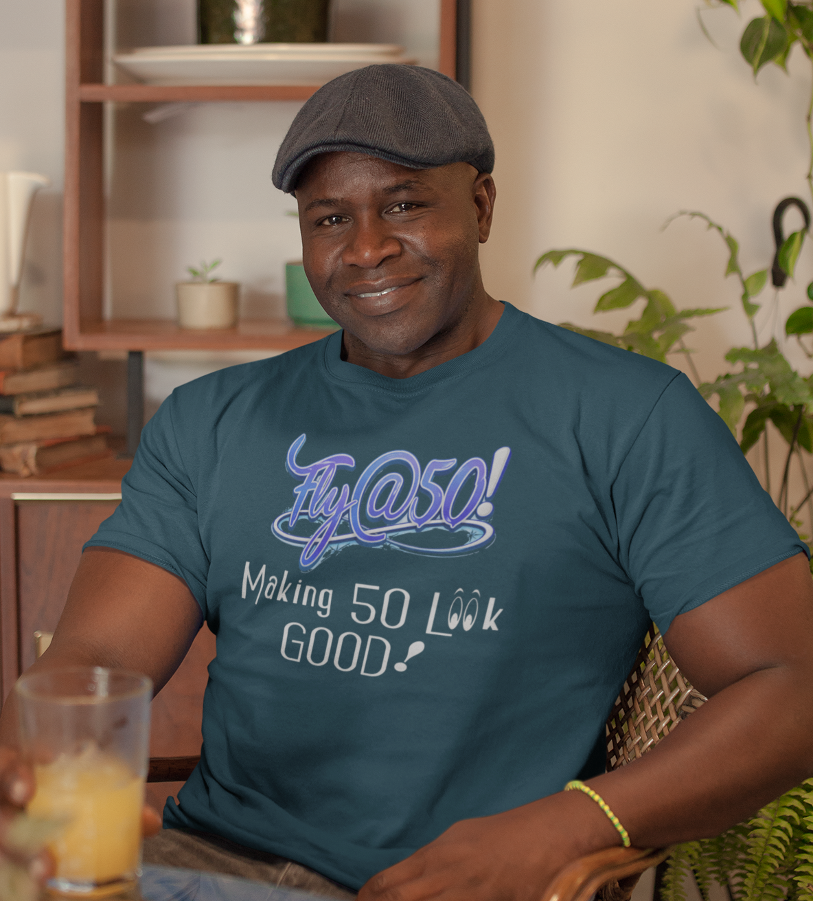 Man wearing a blue t-shirt with text, holding a glass of orange juice in a room with shelves and plants.