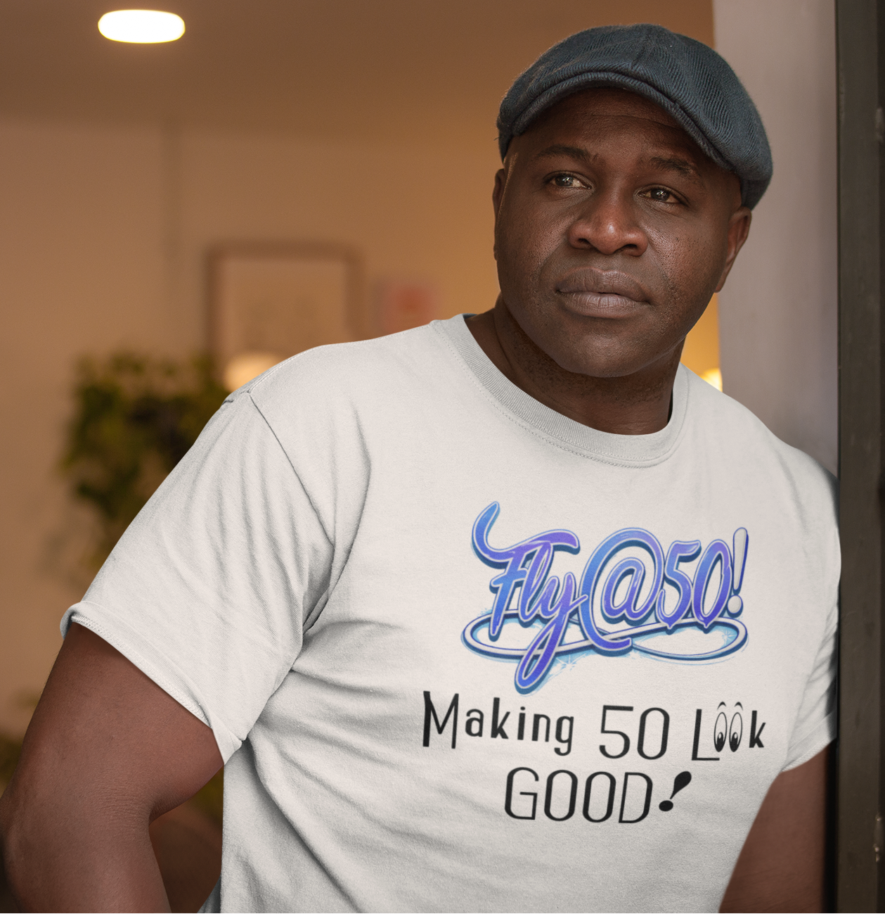 Man wearing a white t-shirt with 'Fly at 50! Making 50 Look Good!' text in a room.