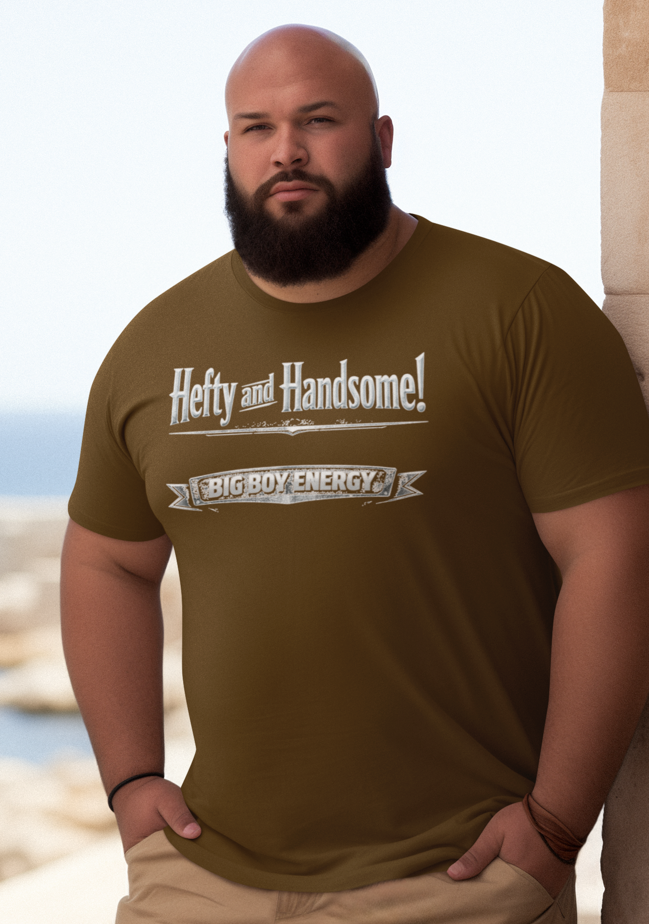 Man wearing a navy blue t-shirt with text saying Hefty and Handsome with BIG Boy Energy underneath, standing by a stone wall with a scenic background.