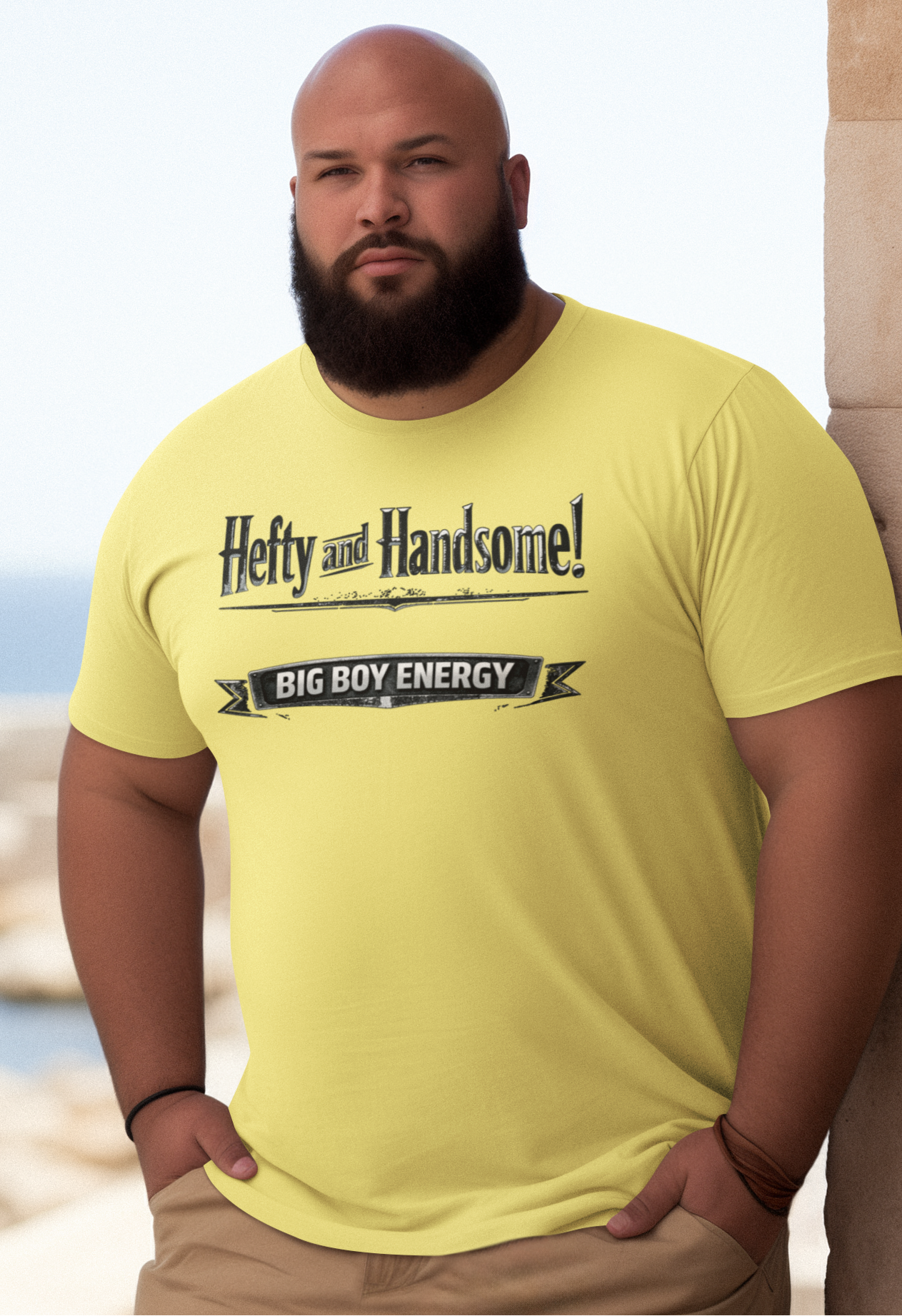 Man wearing a navy blue t-shirt with text saying Hefty and Handsome with BIG Boy Energy underneath, standing by a stone wall with a scenic background.