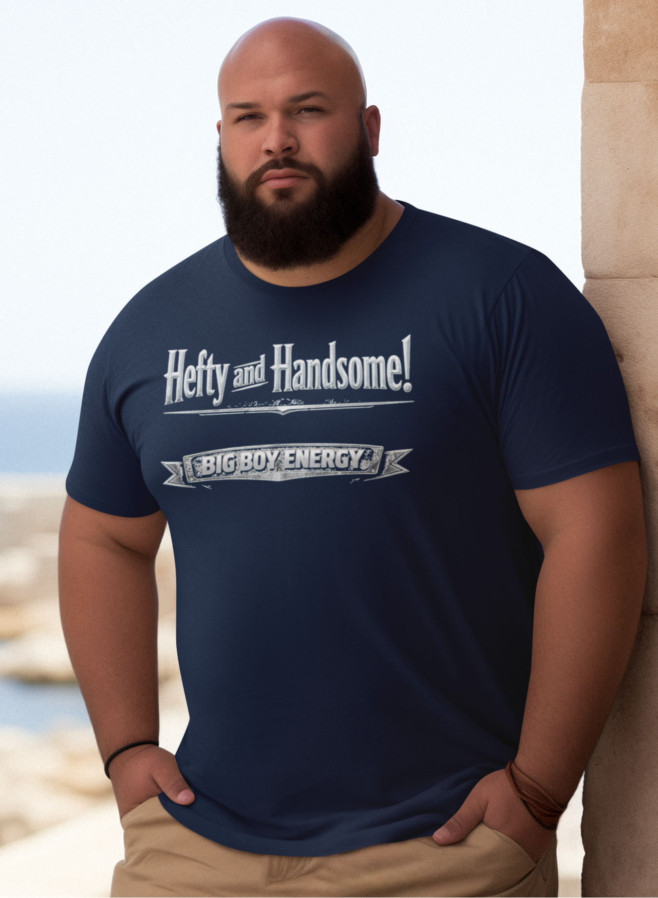 Man wearing a navy blue t-shirt with text saying Hefty and Handsome with BIG Boy Energy underneath, standing by a stone wall with a scenic background.