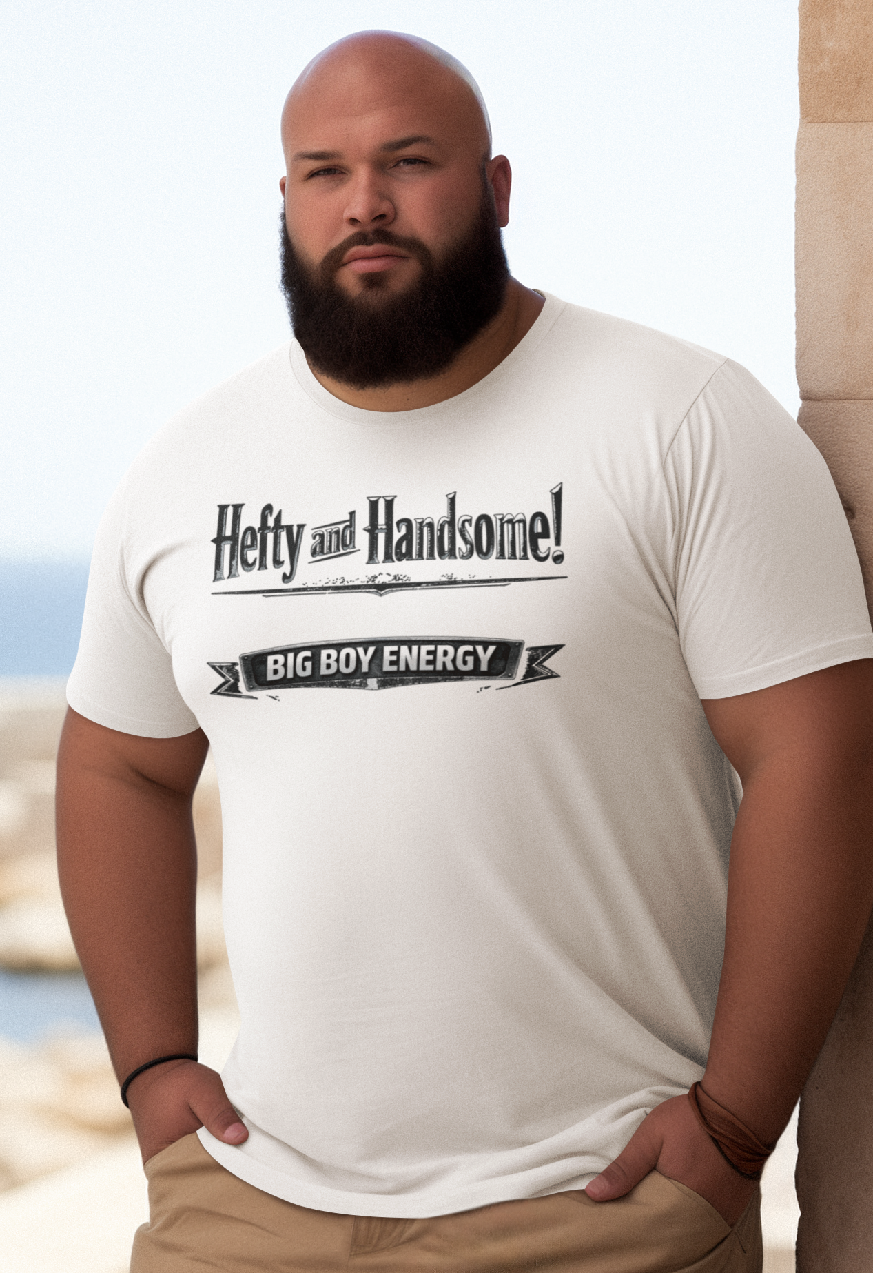 Man wearing a navy blue t-shirt with text saying Hefty and Handsome with BIG Boy Energy underneath, standing by a stone wall with a scenic background.