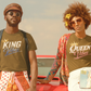 Two people standing in front of a vintage car with a suitcase, wearing matching t-shirts with 'King' and 'Queen' text.